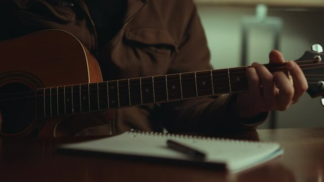 Musician Strums Chords before Hand Writing New Music in Notebook