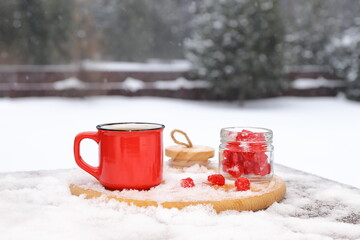 Obraz na płótnie Canvas A red mug of hot tea stands near glass jar with dry cherry berries on a snow-covered wooden tray on the winter forest landscape background. 