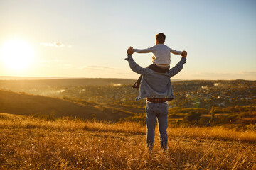 Fathers day. Back view of a little child boy sitting on his fathers shoulders holding hands and looking into the distance enjoying sunset. Father walking with son outdoors. - Powered by Adobe