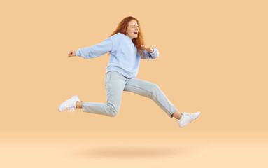 Fototapeta na wymiar Young girl going somewhere in hurry. Full body side profile view happy pretty woman in pastel blue sweatshirt, light gray pants and white shoes jumping and running very fast on beige studio background