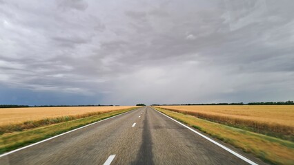 Road passes among fields with ripened wheat against the background of sky with rain clouds