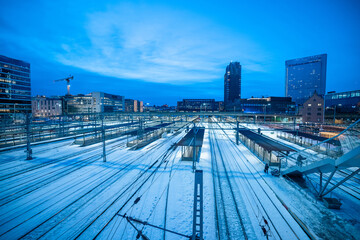 Train station of Oslo seen from one of the overpasses from the bus station. Many tracks seen leading into the station, surrounded by tall buildings. Evening blue hour..