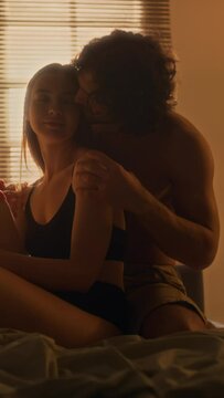 Vertical shot of young affectionate man caressing his girlfriend on bed in cozy bedroom with warm light