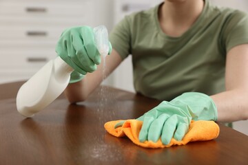 Woman with spray bottle and microfiber cloth cleaning wooden table in room, closeup
