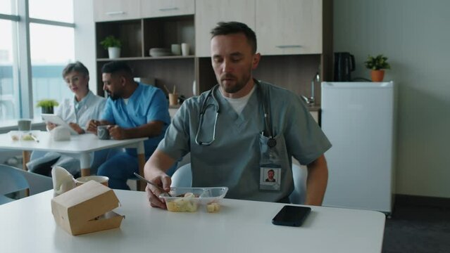 Male hospital worker in scrubs walking with food container in break room, sitting at table and eating his lunch