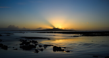 Sunbeams glowing at the crest of Rangitoto Island as the sun is rising. Milford Beach. Auckland.
