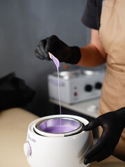 Hot violet wax used for hair removal. Close-up of depilation master hands in black protective gloves holds spatula with melted wax. Device for melting wax