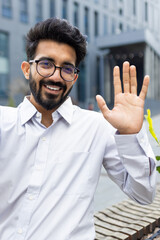 Smiling modern businessman waving hello, showing a friendly gesture, standing outdoors with a cityscape backdrop.
