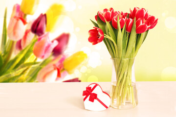 Greeting card template. Decorative bouquet of red tulips and a gift box in shape of a heart on table over blurred background. Spring, valentine, mothers or wedding day.