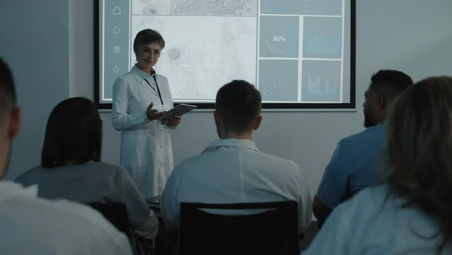 Female scientist in white coat holding digital tablet and giving presentation about microbiology research to healthcare professionals during medical conference