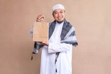 Asian muslim shopaholic man smiling while holding shopping bag on beige background