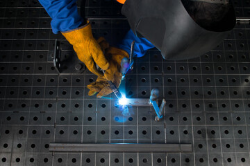 a worker wearing protective gloves and a mask, using a welding machine to fasten..two metal parts,...