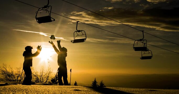 Silhouette of two friends against sunset sky throwing show in the air beneath a row of empty ski lift chairs. Skiers and chairlifts in winter resort. 120fps on cinema camera