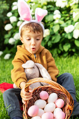 Little caucasian boy wears bunny ears and collects easter eggs to a basket on an easter egg hunt in the garden