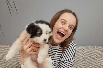 Excited brown haired woman dressed in striped shirt embracing small puppy showing her new small dog to camera exclaiming happily while sitting on sofa at home in living room