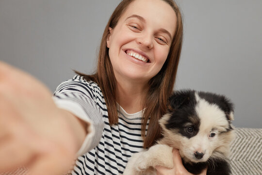 Smiling cheerful overjoyed woman in striped shirt sitting on sofa hugging her black and white puppy making point of view photo captures moment with new pet
