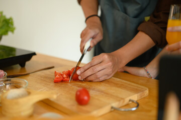 Young man cutting tomato on wooden board at kitchen counter. Closeup view