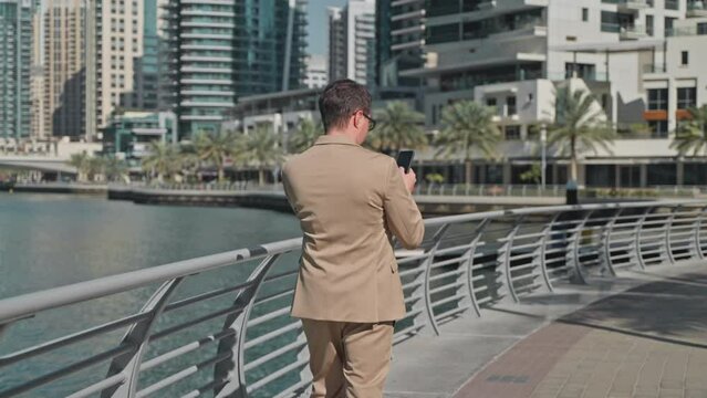 Businessman browsing smartphone walking on waterfront modern megapolis in UAE.