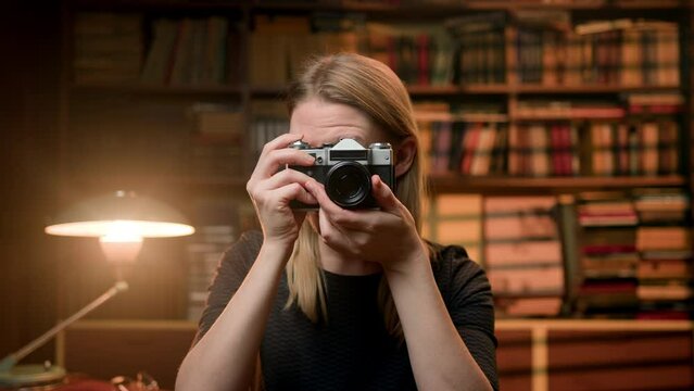 beautiful blonde takes photographs with a retro camera in a library room. A pretty young woman holds a silver camera in her hands. The girl takes pictures and smile