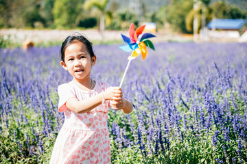 A cheerful little girl stands in a flower garden holding a toy pinwheel. The vibrant springtime evokes joy and flying pinwheels embodying childhood happiness and freedom in nature's sunny embrace.