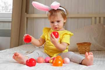 Baby girl with bunny ears. Cute funny baby play Easter eggs at home. Child playing colorful eggs...