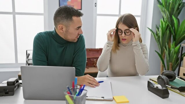 Man and woman converse seriously in a bright office, with laptop, glasses, and stationery on the desk.