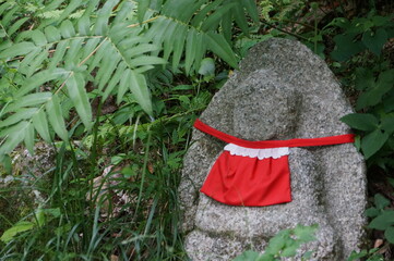 A Japanese stone statue wearing a red hand-stitched bib stands among green trees at Kiyomizu Temple...