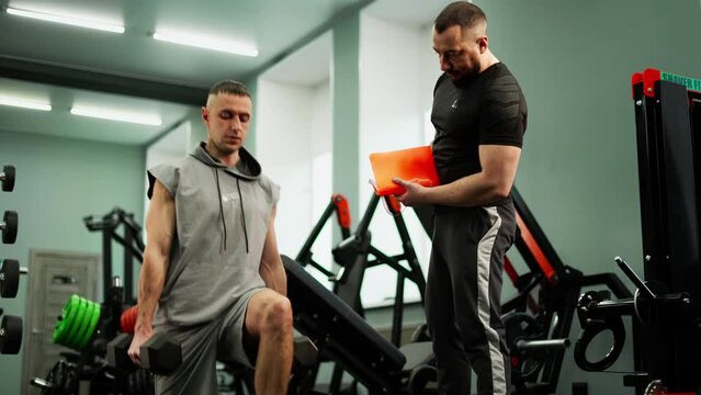 The coach monitors the correctness of the student's exercise with dumbbells in the gym. Bodybuilding classes on special equipment.