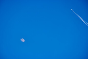 the moon seen during the day in the blue sky and an airplane that leaves traces behind it.