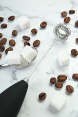 Black milk frother wand, sugar cubes and coffee beans on white marble table, closeup