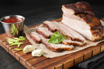 Pieces of baked pork belly served with sauce and parsley on black textured table, closeup