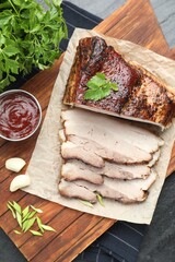 Pieces of baked pork belly served with sauce and parsley on black textured table, top view