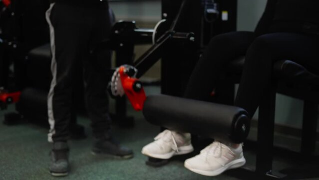 A female athlete performs a leg exercise in a special simulator under the supervision of a coach. The trainer corrects and monitors the correctness of the exercise.