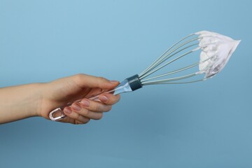 Woman holding whisk with whipped cream on light blue background, closeup