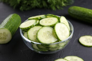 Cut cucumber in glass bowl and fresh vegetables on dark gray textured table, closeup