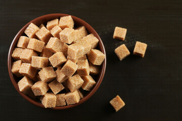 Brown sugar cubes in bowl on black wooden table, top view