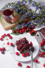 Chocolate cake with sweet cherry, cup of tea on vintage book and branch of blue flowers on white background..