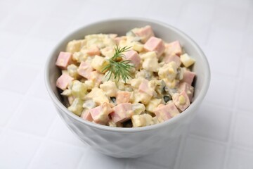 Tasty Olivier salad with boiled sausage in bowl on white tiled table, closeup