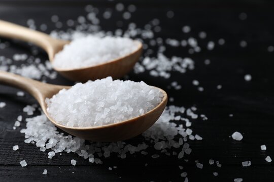 Organic salt in spoons on black wooden table, closeup. Space for text