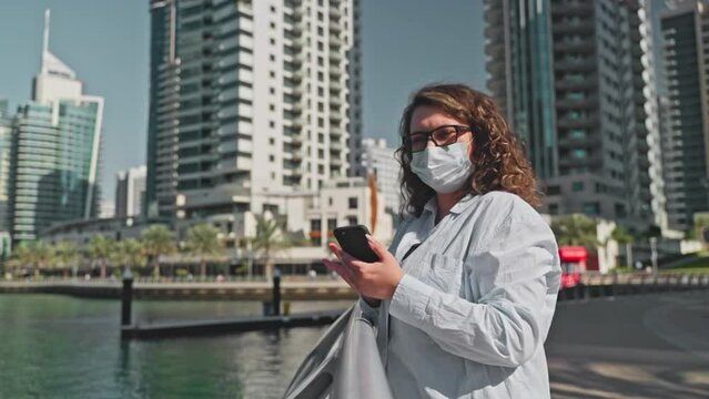 Tourist woman in medical face mask browsing phone walking on waterfront in city.