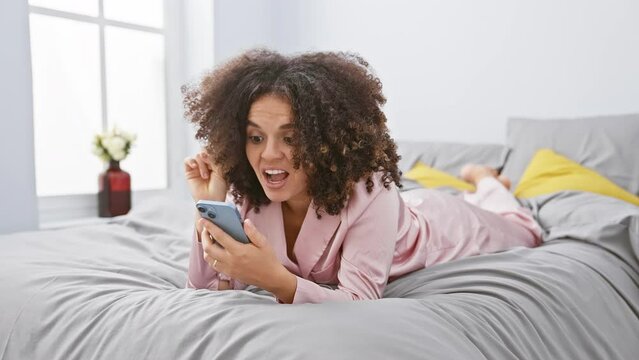 Hispanic curly hair woman lying on bed using smartphone smiling with an idea or question pointing finger with happy face, number one at bedroom