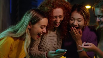 Young group of gen z people having fun surprised using cell phone together outside watching something funny at mobile. Cheerful community of student enjoying social media content. Multiethnic friends