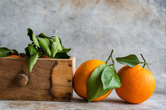 Ripe oranges with fresh green leaves attached are displayed, some resting on a textured surface and others nestled in a wooden crate, evoking a sense of fresh harvest