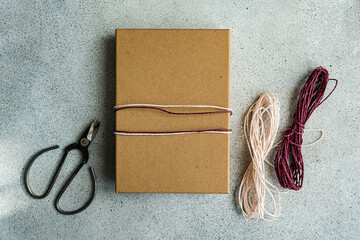 A simple yet elegant gift wrapped in brown paper with a pink and white twine, alongside scissors and additional twine on a textured grey background