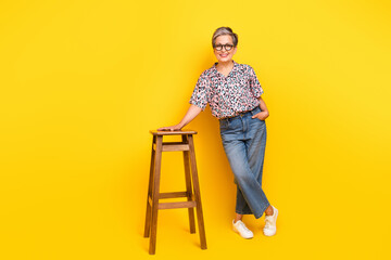 Full body photo of attractive pensioner woman stand lean on chair dressed stylish leopard print clothes isolated on yellow color background
