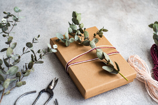 A beautifully wrapped gift in brown paper adorned with eucalyptus branches and twine, next to scissors and extra twine on a concrete background