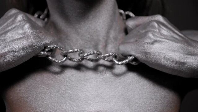 Close-up of a slender woman with bare shoulders with silver body art on her skin posing from a studio with a large chain around her neck, she clutches the chain with her hands and tears it.