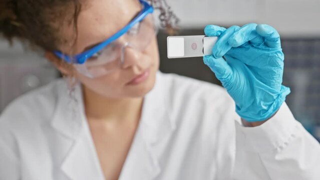 A focused hispanic woman with curly hair examines a slide in a laboratory, wearing glasses and gloves for safety.