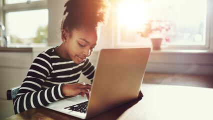 Little young black girl sits at the kitchen table and writes on a laptop - 740677234