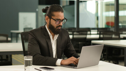 Busy concentrated Indian male employer company corporate Arabian entrepreneur boss CEO business man Muslim businessman at office with laptop working typing thinking solution idea pensive looking away - Powered by Adobe